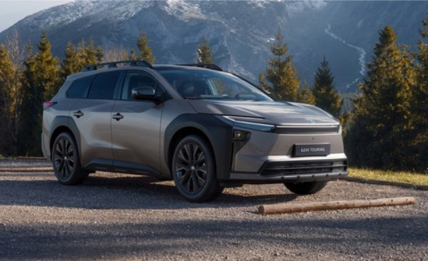 A grey SUV parked on a gravel surface with trees and mountains in the background.