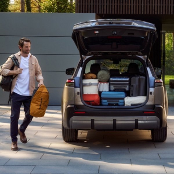 Car boot loaded with luggage and items, man with a bag walking towards the car.