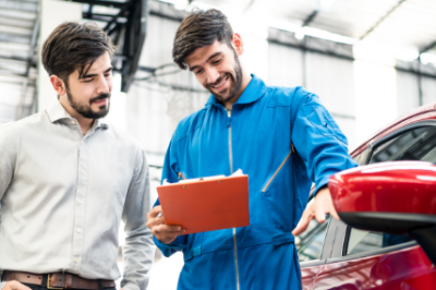 Mechanic discussing car details with a customer, holding a clipboard next to a red car.