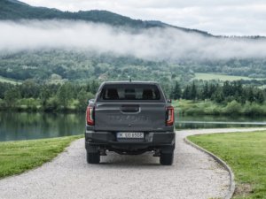 A grey pickup truck parked on a gravel path by a lake and mountains.