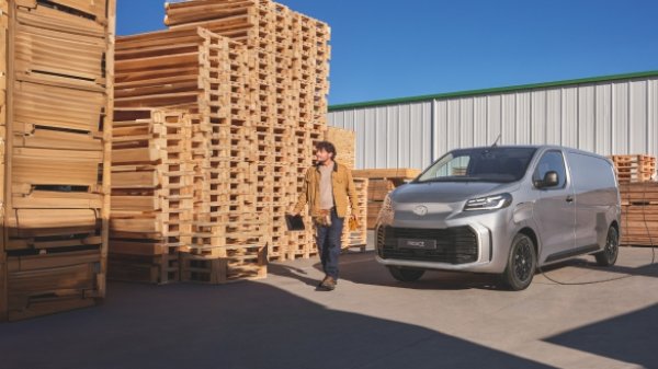 Man walking beside a parked silver van and a stack of wooden pallets.