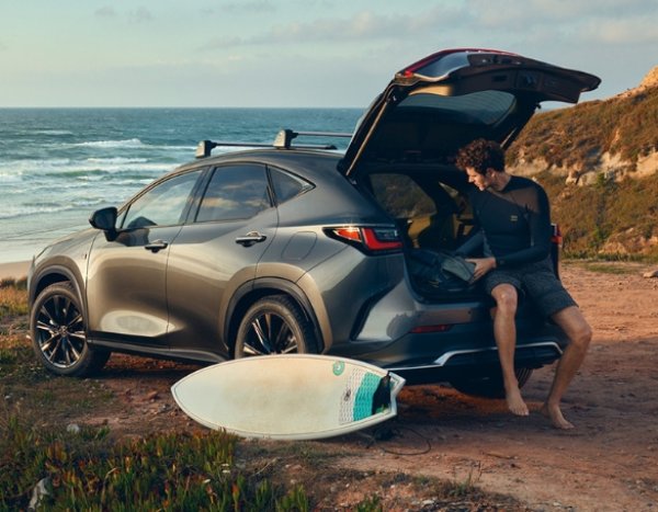 Man sitting in SUV boot by the sea with a surfboard on the ground nearby.