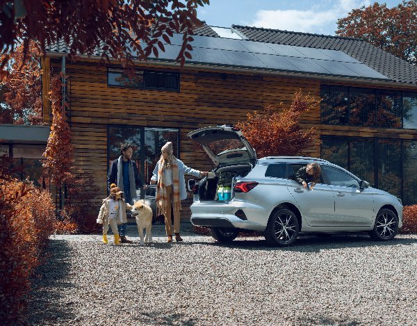 Family with a dog unloading luggage from a white car in front of a modern house.