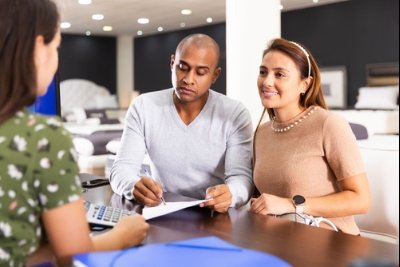 A couple sits at a table, looking at documents with a salesperson in a shop.