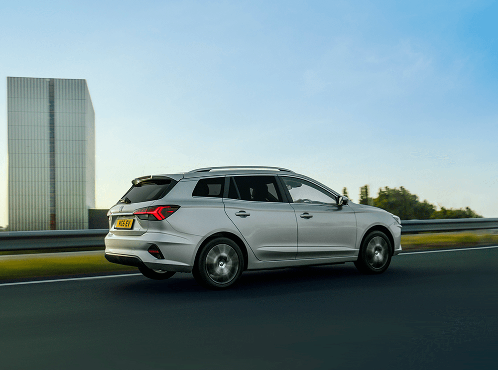 A silver estate car driving on a highway beside a large, modern building.