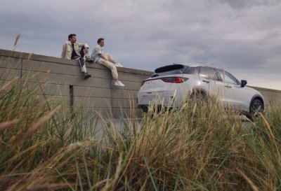 Three people sitting on a wall near a silver SUV with tall grass in the foreground.